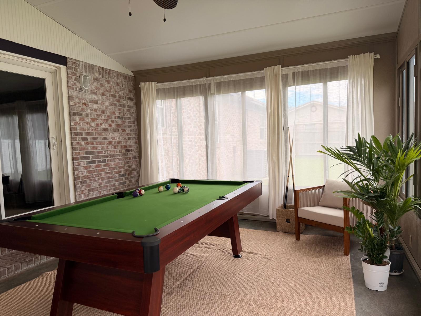 Pool table in a bright sunroom with plants and a woven chair